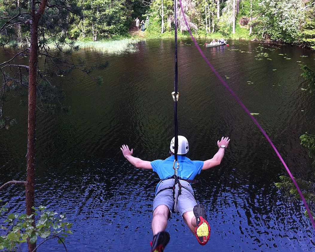 En person med hvit hjelm og blå skjorte som kjører zipline over en rolig innsjø omgitt av grønne trær.