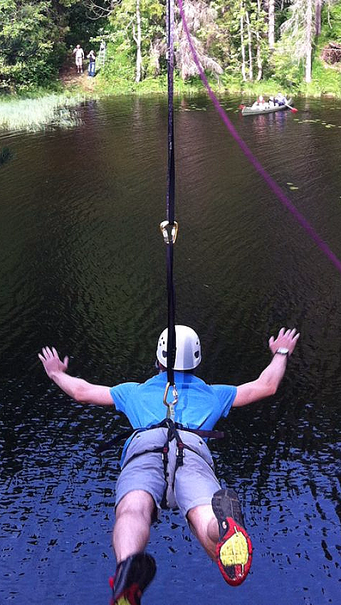 En person med hvit hjelm og blå skjorte som kjører zipline over en rolig innsjø omgitt av grønne trær.