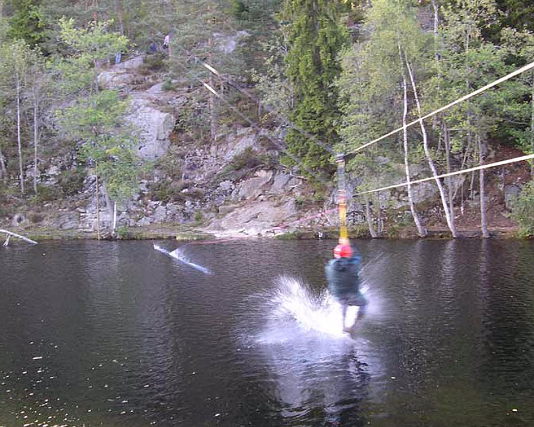 En person med rød hjelm kjører zipline over vannet og skaper en vannsprut, med skog og klipper i bakgrunnen.