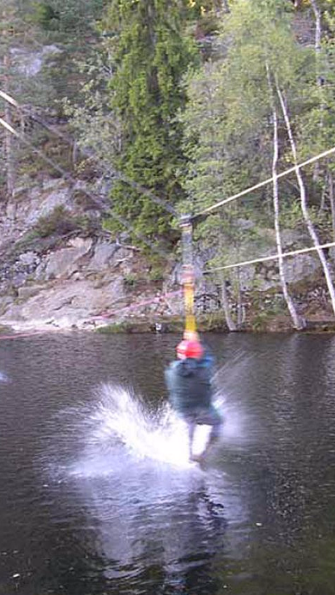 En person med rød hjelm kjører zipline over vannet og skaper en vannsprut, med skog og klipper i bakgrunnen.