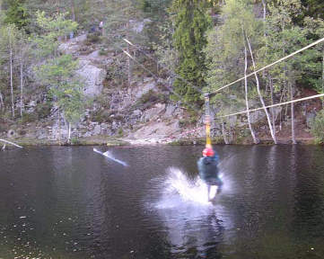 En person med rød hjelm kjører zipline over vannet og skaper en vannsprut, med skog og klipper i bakgrunnen.