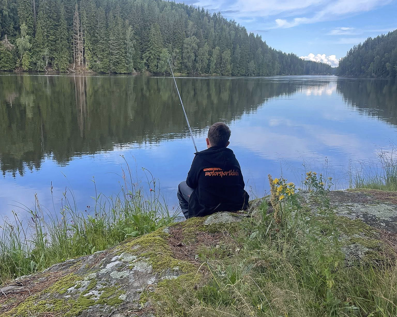 En person sitter på en mosegrodd stein ved en rolig innsjø og fisker, med skog og blå himmel som speiler seg i vannet.