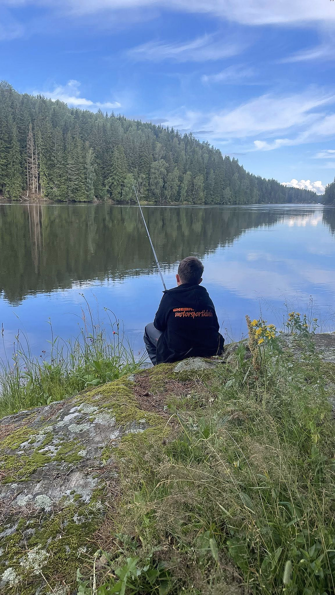 En person sitter på en mosegrodd stein ved en rolig innsjø og fisker, med skog og blå himmel som speiler seg i vannet.