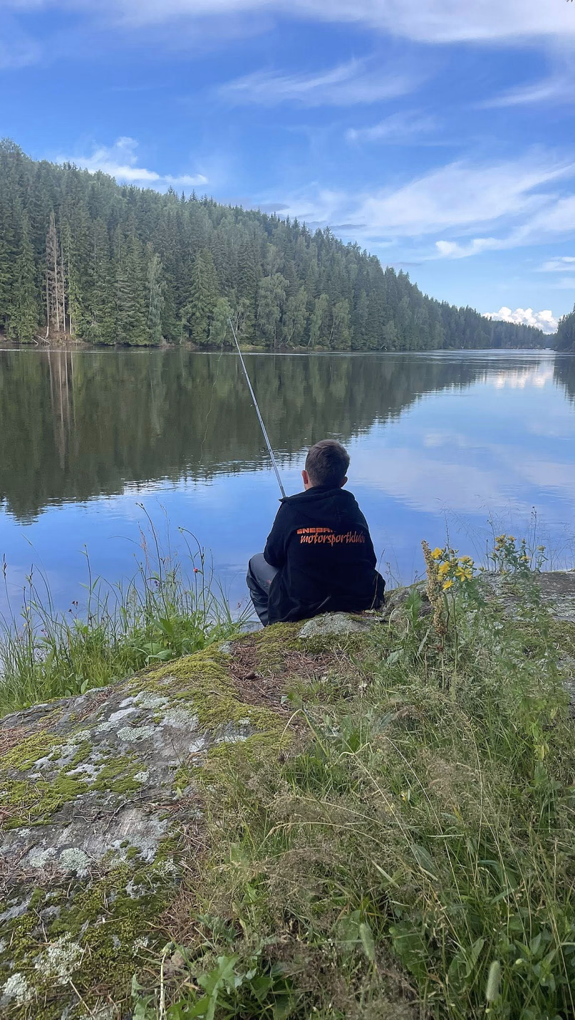 En person sitter på en mosegrodd stein ved en rolig innsjø og fisker, med skog og blå himmel som speiler seg i vannet.