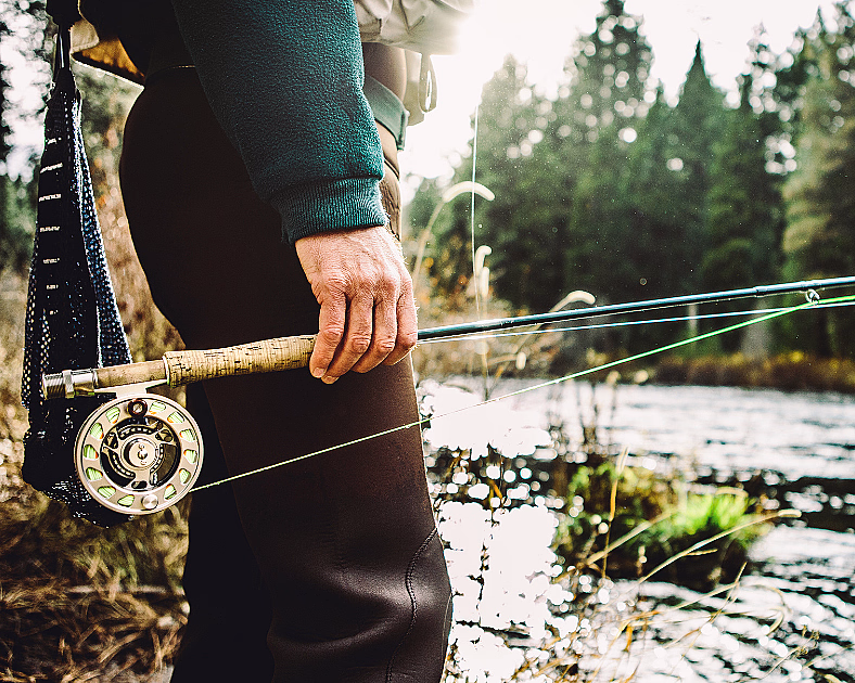 En person i grønn genser holder en fiskestang ved en elv med skog og sollys i bakgrunnen.