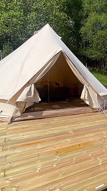 Et beige lerretstelt på en treterrasse med to grønne fluktstoler og et lite bord, omgitt av gress og trær under en blå himmel.