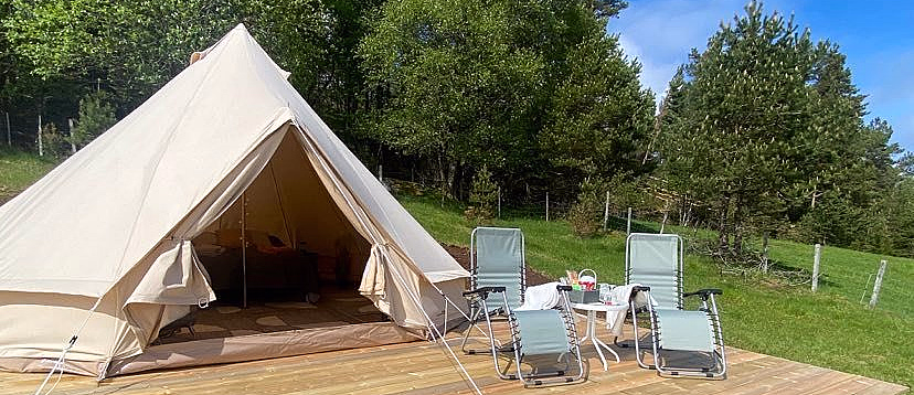 Et beige lerretstelt på en treterrasse med to grønne fluktstoler og et lite bord, omgitt av gress og trær under en blå himmel.