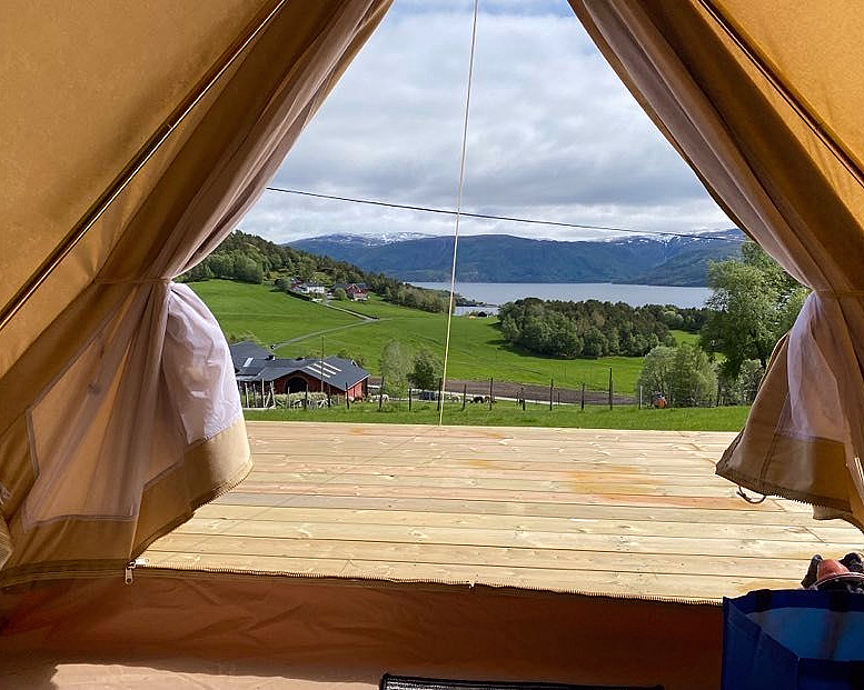 Utsikt fra åpningen av et telt mot en treterrasse, en innsjø og snødekte fjell i det fjerne.