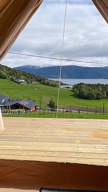 Utsikt fra åpningen av et telt mot en treterrasse, en innsjø og snødekte fjell i det fjerne.