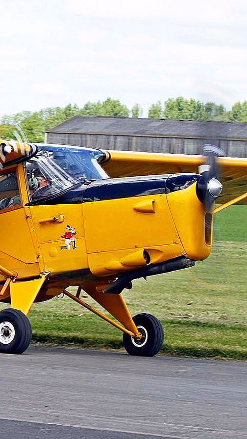 Et gult veteranfly med én propell på en rullebane, med et rødt lønneblad-symbol og tallet 693 på siden, mot en bakgrunn av grønt landskap og en delvis skyet himmel.