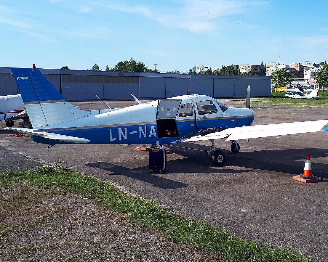 Et lite blått og hvitt småfly med registreringsnummer LN-NA står parkert på en asfaltflate med døren åpen under en klar blå himmel.