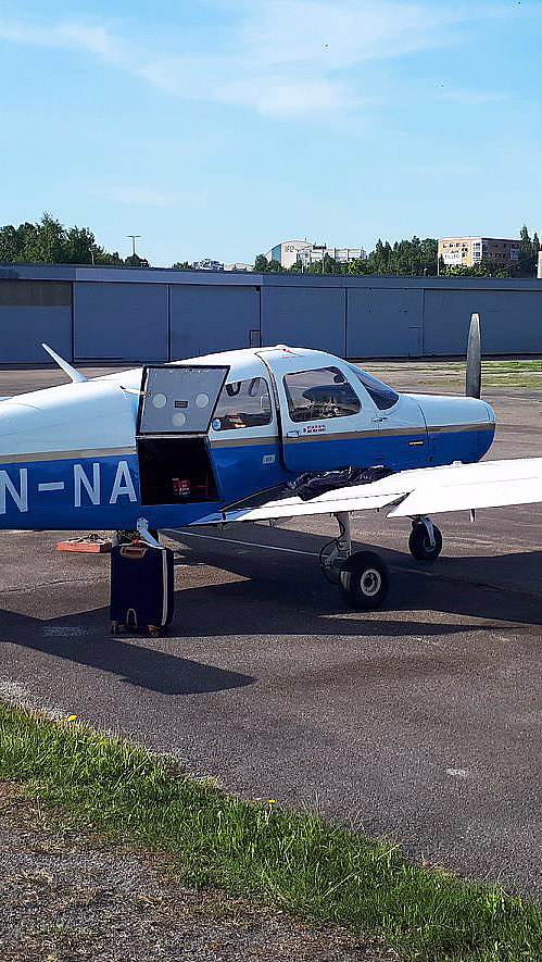 Et lite blått og hvitt småfly med registreringsnummer LN-NA står parkert på en asfaltflate med døren åpen under en klar blå himmel.