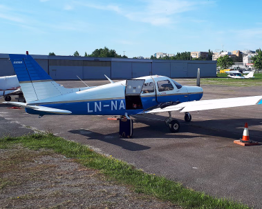 Et lite blått og hvitt småfly med registreringsnummer LN-NA står parkert på en asfaltflate med døren åpen under en klar blå himmel.