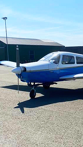 Et lite blått og hvitt småfly med kjennetegnet LN-NAR parkert på en asfaltflate foran to grønne hangarer under en klar blå himmel.