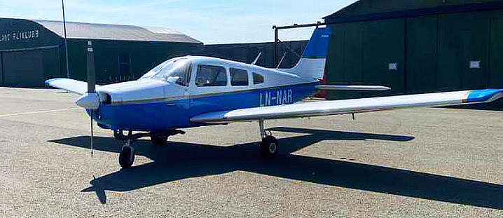 Et lite blått og hvitt småfly med kjennetegnet LN-NAR parkert på en asfaltflate foran to grønne hangarer under en klar blå himmel.
