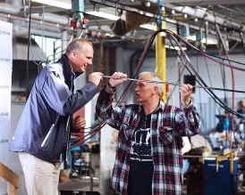 En mann blåser i et langt rør for å forme glass i et glassverksted mens en annen mann ser på