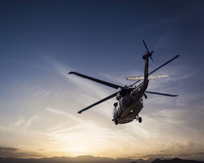 Helikopter i luften sett nedenfra mot en solnedgang og fjellkjede