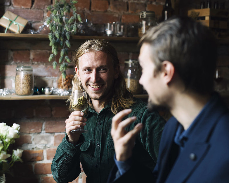 Smilende mann som holder et glass med humle i samtale med andre foran en murvegg