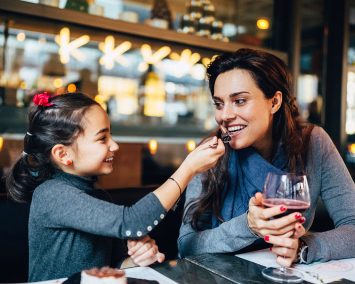 En jente mater en kvinne med en skje ved et restaurantbord
