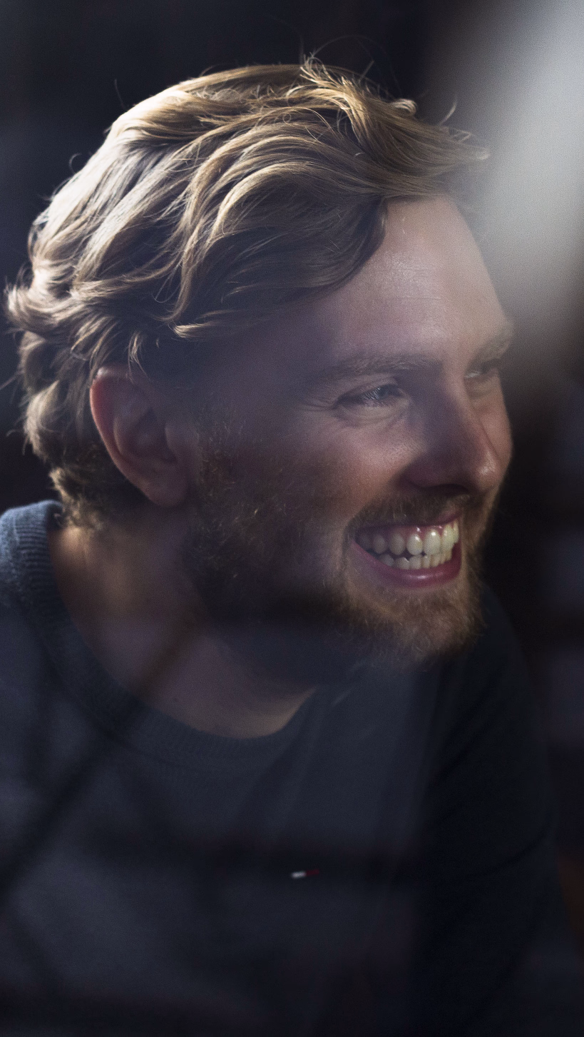 Smilende mann med skjegg sett fra siden i et mørkt lokale