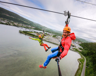 To personer i klatresele som kjører zipline over vann og landskap