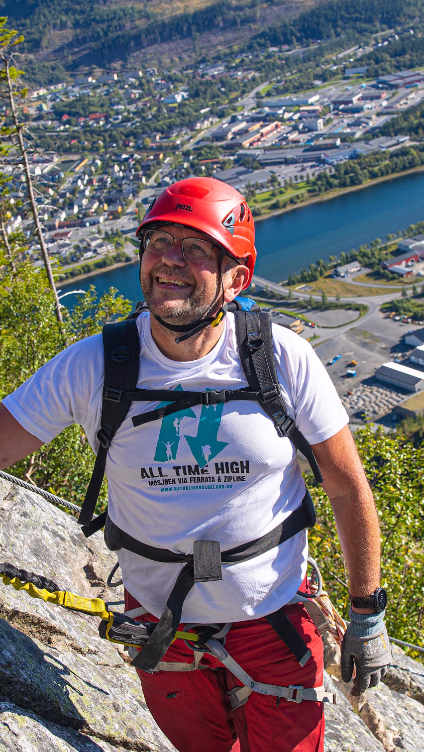 Mann i klatresele og rød hjelm klatrer via ferrata med utsikt over en by og elv
