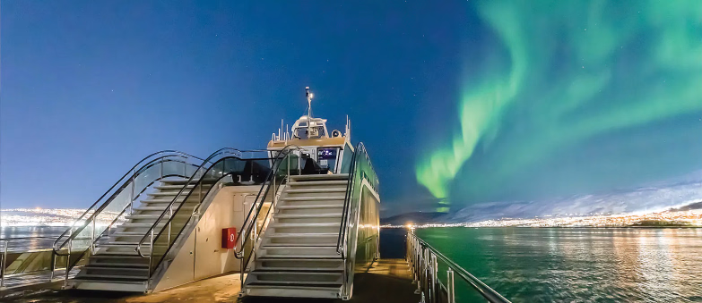 Grønt nordlys på himmelen over en båt ved en opplyst by om natten