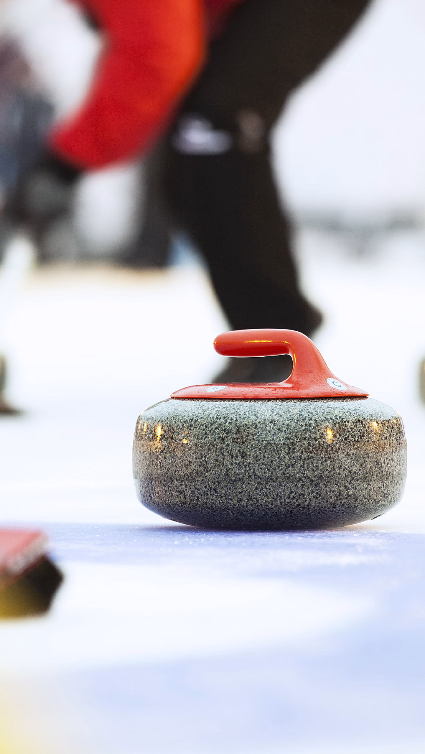 En grå curlingstein med rødt håndtak på isen, med en person i rød jakke som feier med en rød kost i bakgrunnen.