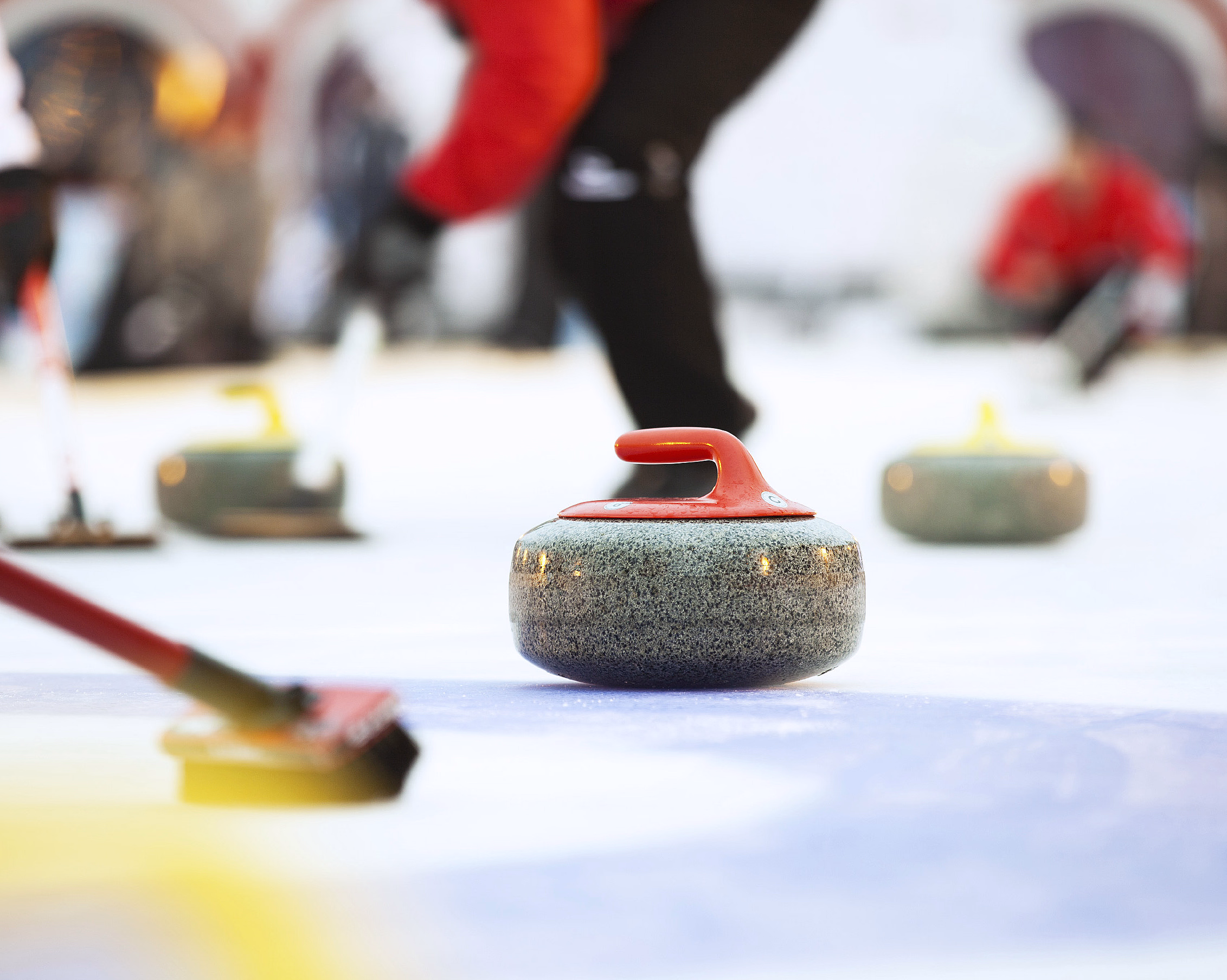 En curlingstein med rødt håndtak på isen, med en person som koster og andre steiner i bakgrunnen.