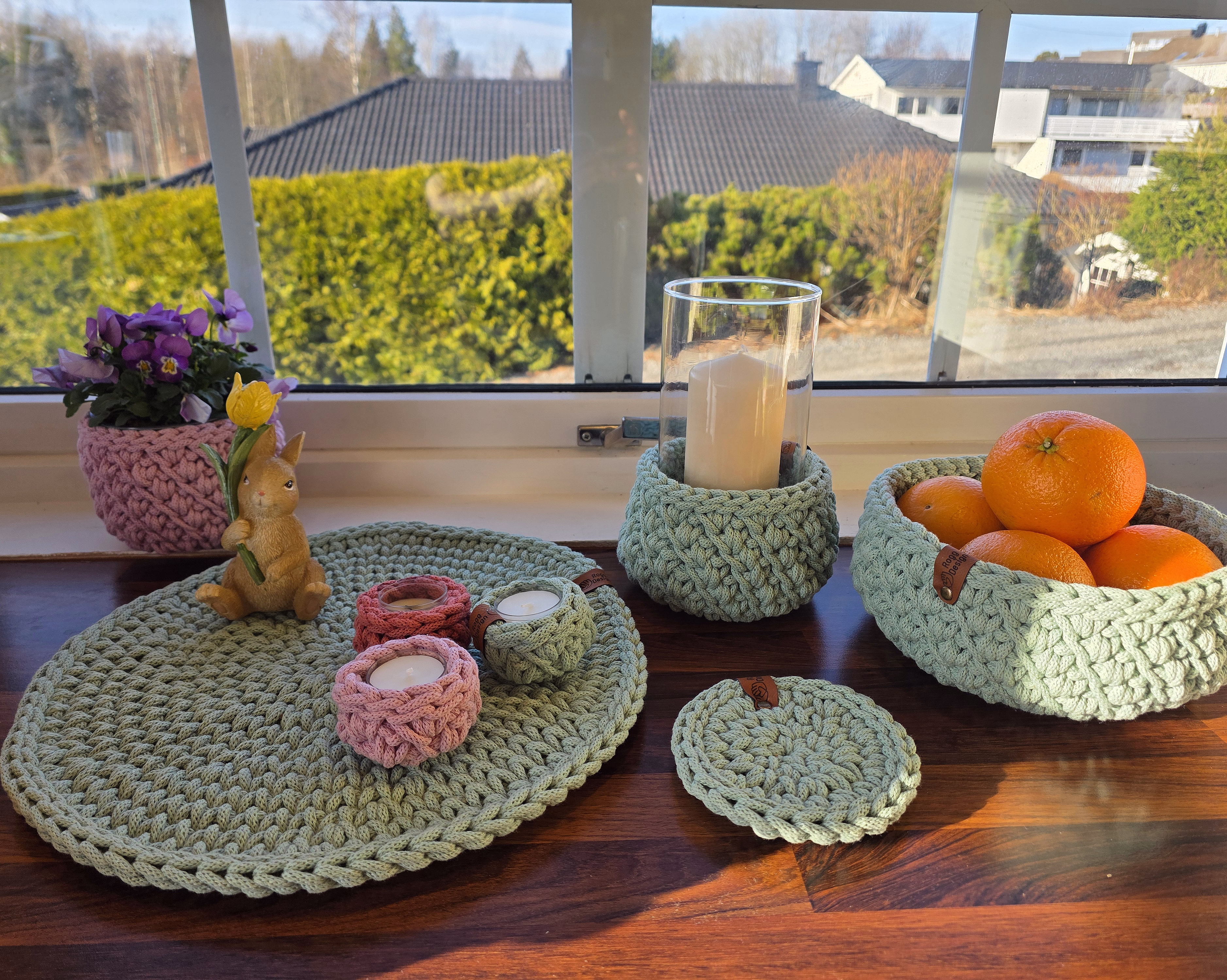Solfylt vinduskarm i mørkt tre med grovheklet interiør i mintgrønt og rosa, inkludert kurver med appelsiner, telysholdere, en blomsterpotte med lilla blomster og en liten harefigur.