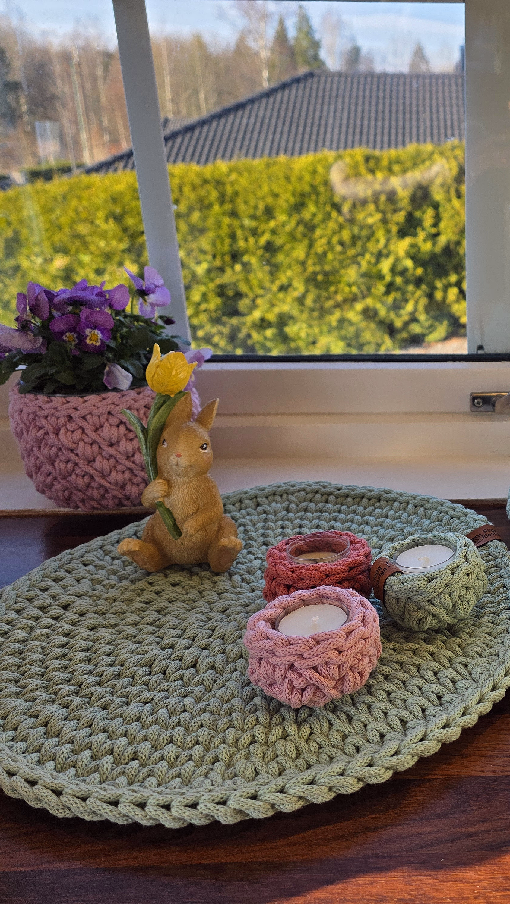 Solfylt vinduskarm i mørkt tre med grovheklet interiør i mintgrønt og rosa, inkludert kurver med appelsiner, telysholdere, en blomsterpotte med lilla blomster og en liten harefigur.