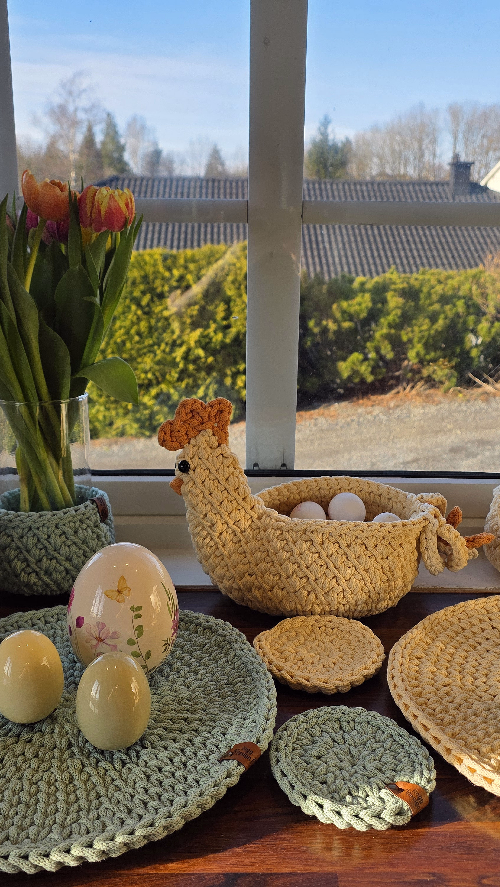 Solfylt vinduskarm pyntet til påske med en strikket hønekurv med egg, gule påskeliljer, rosa tulipaner og små harefigurer på flettede brikker.