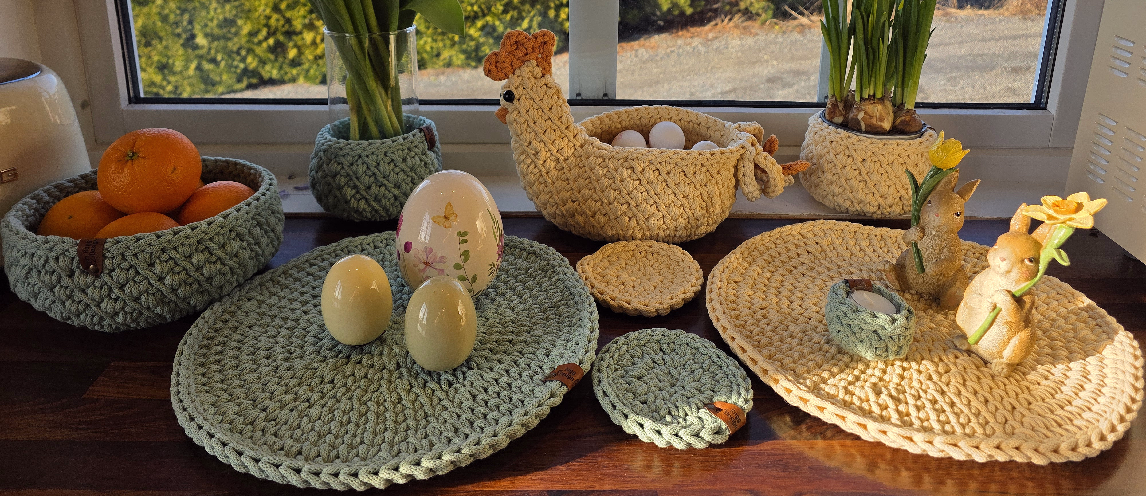 Solfylt vinduskarm pyntet til påske med en strikket hønekurv med egg, gule påskeliljer, rosa tulipaner og små harefigurer på flettede brikker.