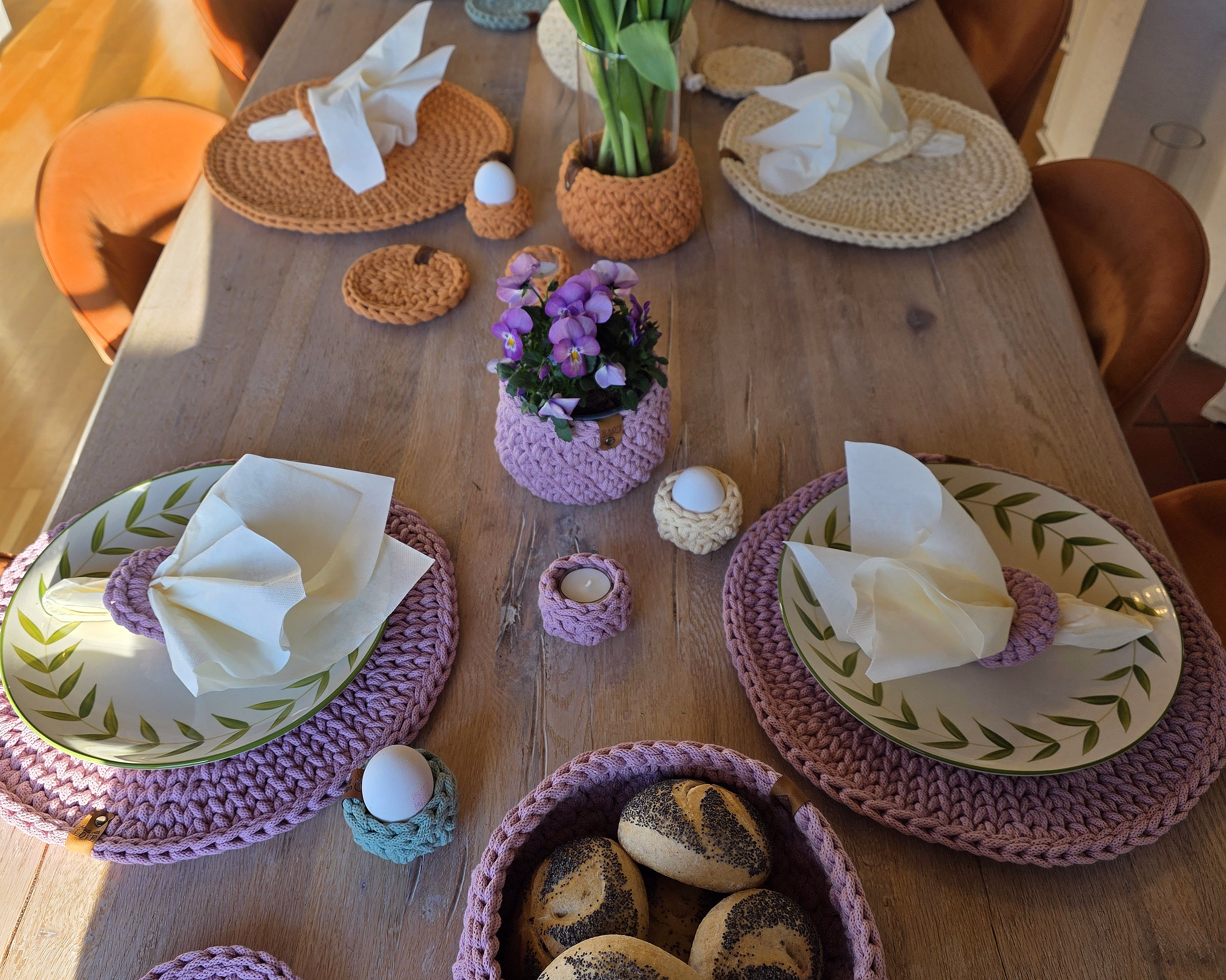 Et langt trebord dekket til et måltid med fargerike heklede brikker, tallerkener med bladkant, oransje tulipaner i en glassvase og en rosa heklet kurv med rundstykker i varmt sollys.