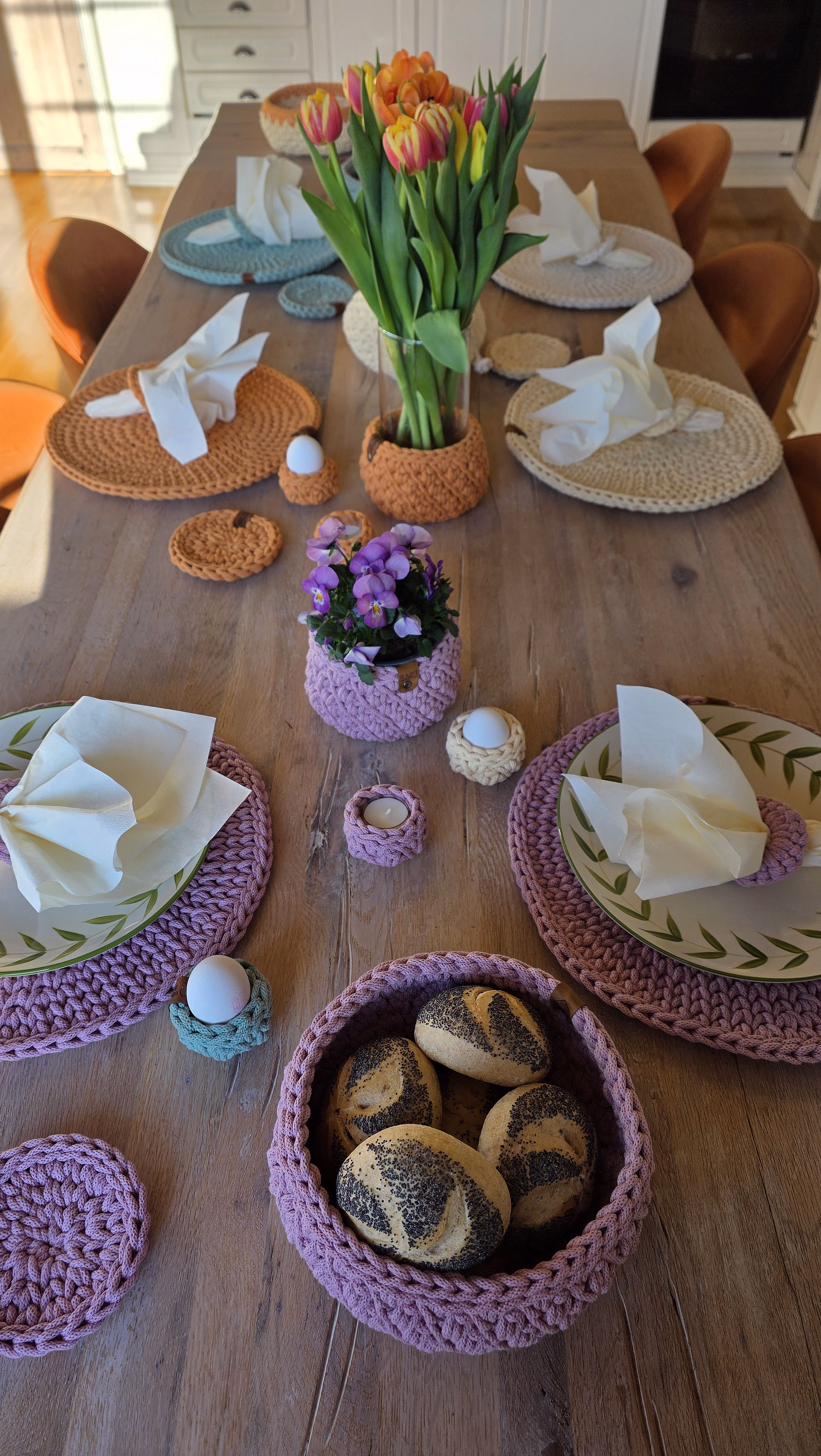 Et langt trebord dekket til et måltid med fargerike heklede brikker, tallerkener med bladkant, oransje tulipaner i en glassvase og en rosa heklet kurv med rundstykker i varmt sollys.
