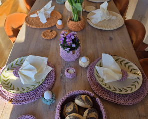 Et langt trebord dekket til et måltid med fargerike heklede brikker, tallerkener med bladkant, oransje tulipaner i en glassvase og en rosa heklet kurv med rundstykker i varmt sollys.
