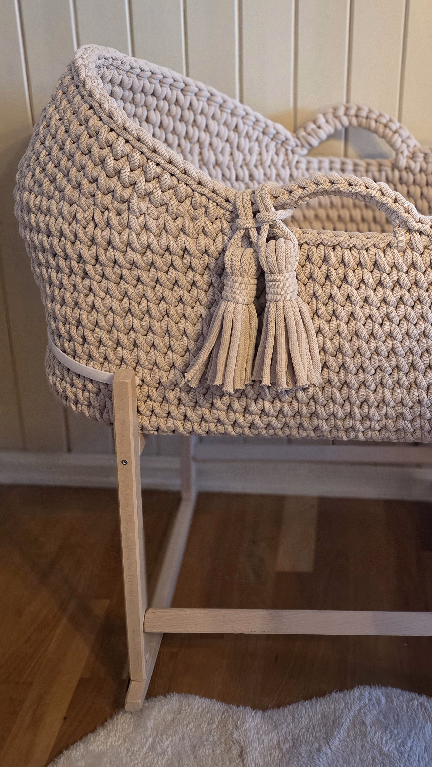 Beige moseskurv i tykk strikk med dusker, plassert på et lyst trestativ foran en lys panelvegg.