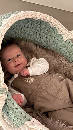 En smilende baby som ligger i en oval, flettet vugge av tykt mintgrønt og kremfarget garn. Vuggen har et mykt grått fôr og står på et hvitt trestativ på et lyst gulv.