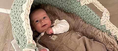 En smilende baby som ligger i en oval, flettet vugge av tykt mintgrønt og kremfarget garn. Vuggen har et mykt grått fôr og står på et hvitt trestativ på et lyst gulv.