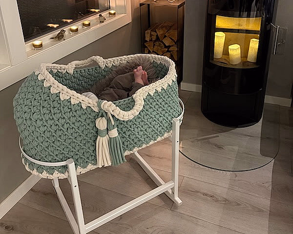 Koselig rom med en mintgrønn og hvit strikket babykurv på stativ, små babyføtter under et grått teppe, og en svart ovn med levende lys i bakgrunnen.