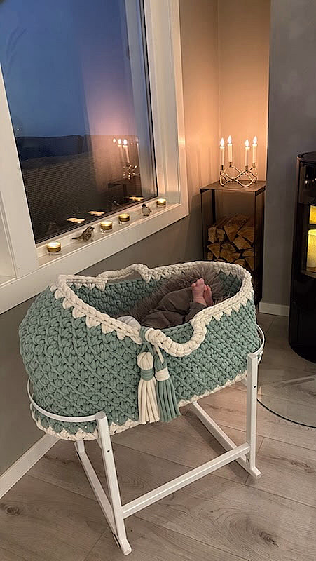 Koselig rom med en mintgrønn og hvit strikket babykurv på stativ, små babyføtter under et grått teppe, og en svart ovn med levende lys i bakgrunnen.
