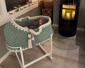 Koselig rom med en mintgrønn og hvit strikket babykurv på stativ, små babyføtter under et grått teppe, og en svart ovn med levende lys i bakgrunnen.