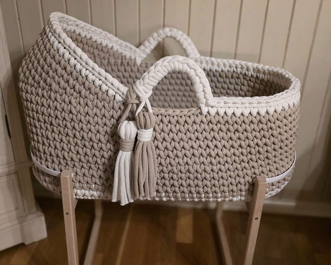 Oval babykurv i beige tykkstrikk med hvit kant og dusker, plassert på et lyst trestativ.