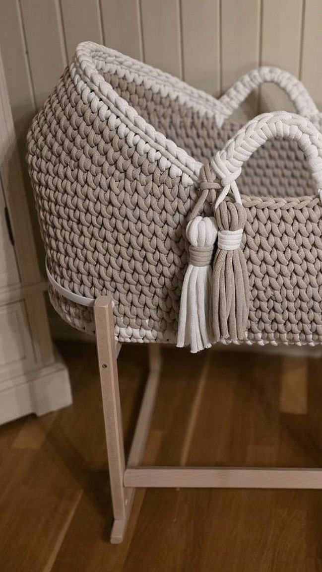 Oval babykurv i beige tykkstrikk med hvit kant og dusker, plassert på et lyst trestativ.