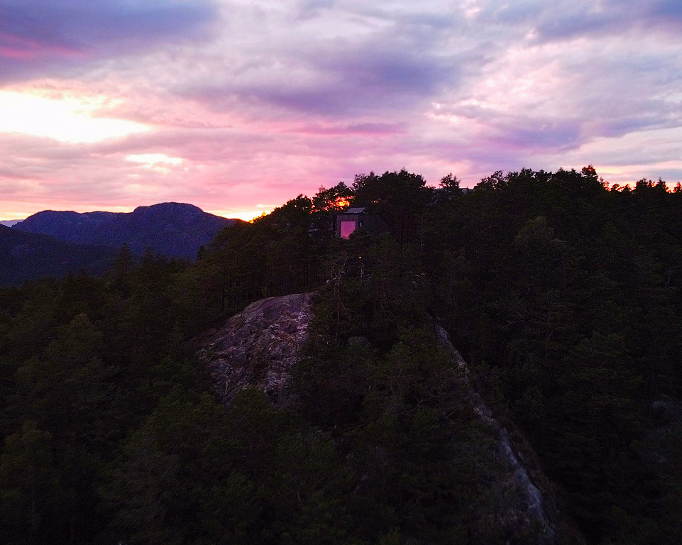 En liten mørk hytte på en steinete åsrygg omgitt av furuskog, med en fargerik solnedgang i lilla, rosa og oransje. Et vindu i hytta lyser opp i rosa, med fjerne fjell i bakgrunnen.