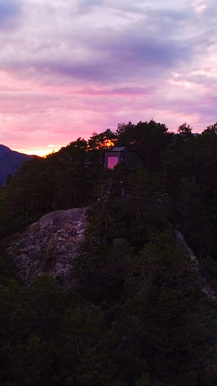 En liten mørk hytte på en steinete åsrygg omgitt av furuskog, med en fargerik solnedgang i lilla, rosa og oransje. Et vindu i hytta lyser opp i rosa, med fjerne fjell i bakgrunnen.