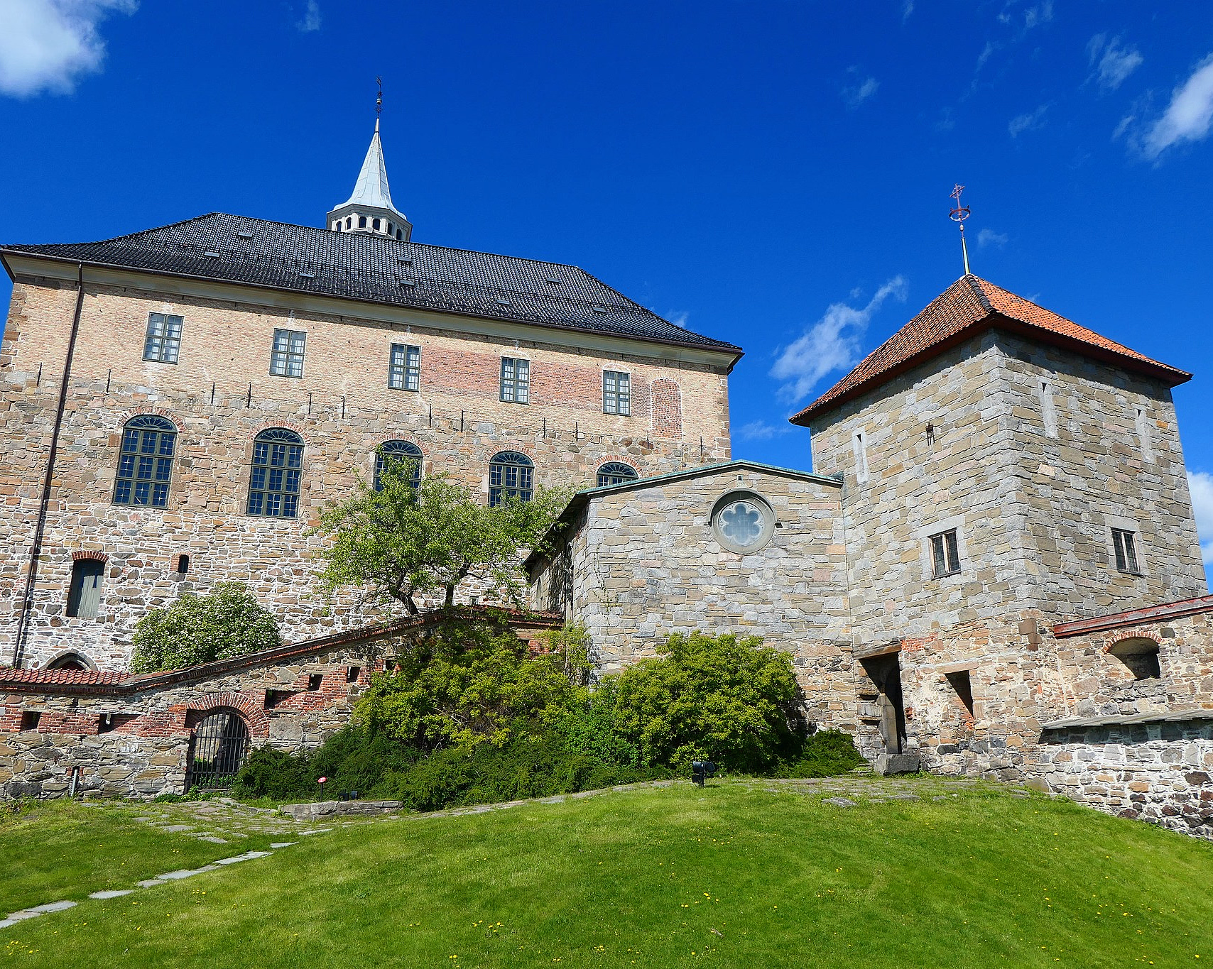 Et stort steinslott i grå og brun stein med et hvitt spir, et firkantet tårn med rødt tak og buede vinduer på en grønn gressbakke under en blå himmel.