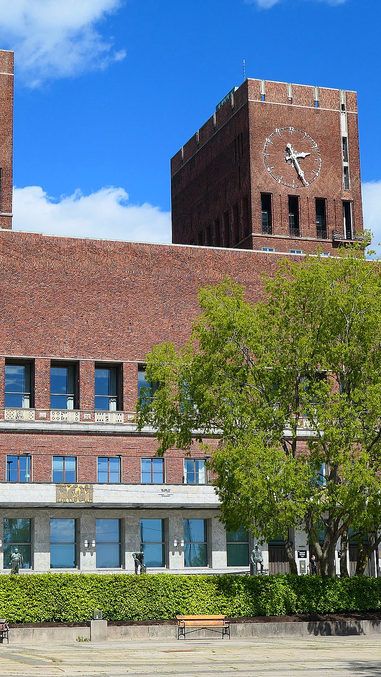 Stor rød mursteinsbygning med to høye tårn, der det høyre tårnet har en stor klokke. Foran er en steinbelagt plass med statuer og grøntarealer under en blå himmel.