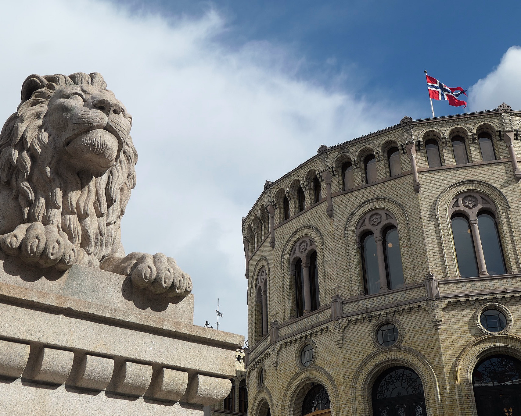 Stor løvestatue i stein foran en rund, gul mursteinsbygning med det norske flagget på taket under en blå himmel.