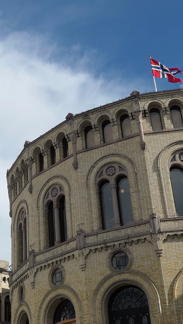 Stor løvestatue i stein foran en rund, gul mursteinsbygning med det norske flagget på taket under en blå himmel.